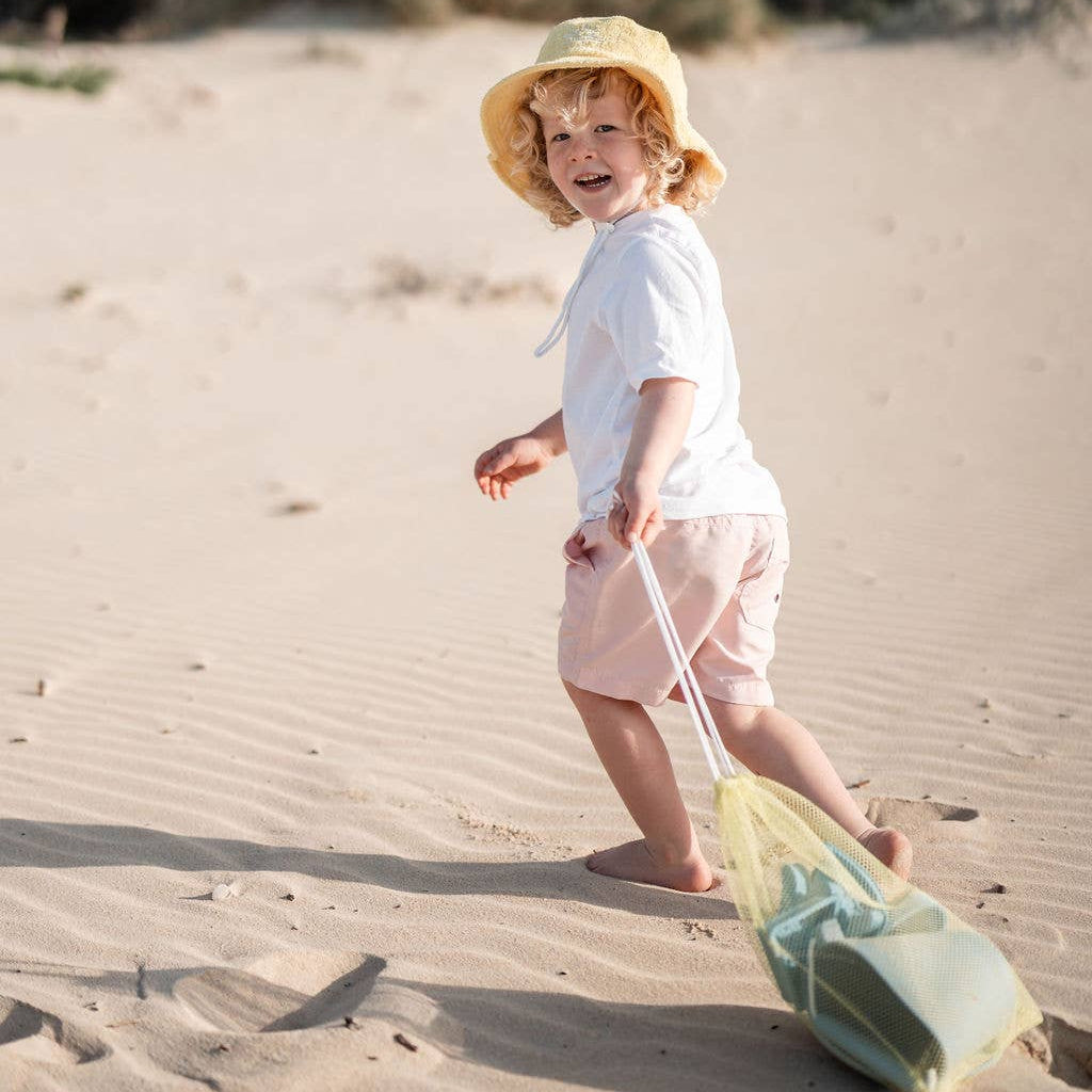 Child on a sandy beach wearing a hat and holding a mesh toy bag